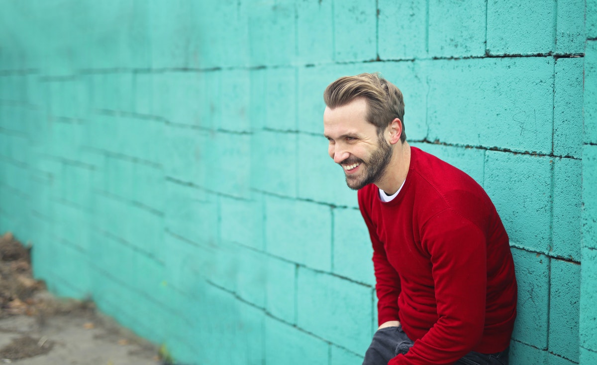 A photo of a joyful man wearing a red-colored sweater leaning on an evergreen wall as a symbol of sound mental health.